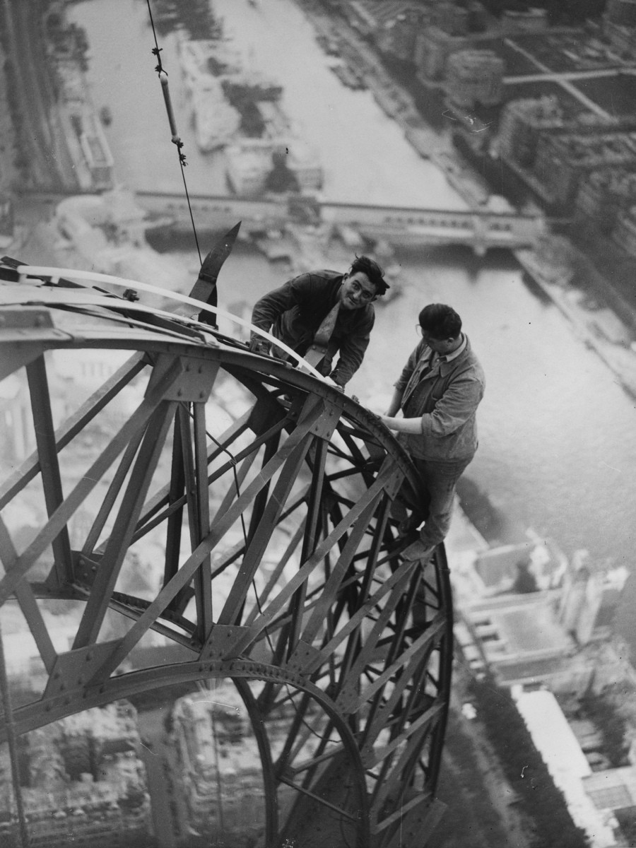 High above the River Seine two electricians work on the lights on the Eiffel Tower which will illuminate the Paris Exhibition at night.